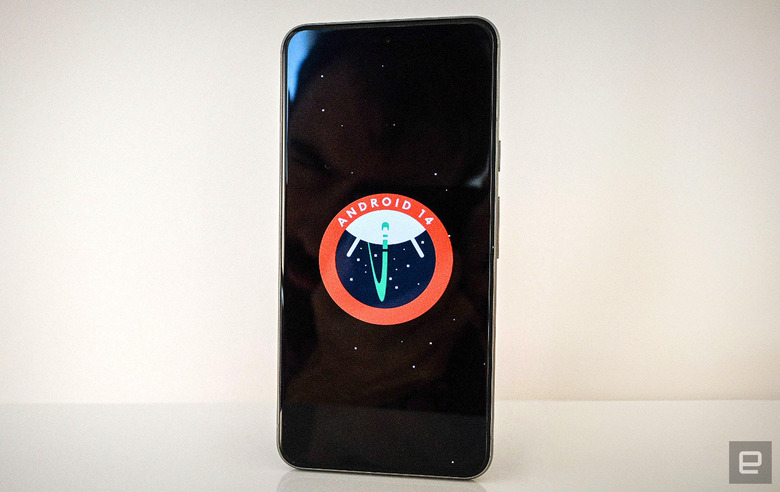 A photo of a Pixel 8 with the Android 14 logo displayed on the screen. The phone stands upright on a white shelf, against a white wall.   