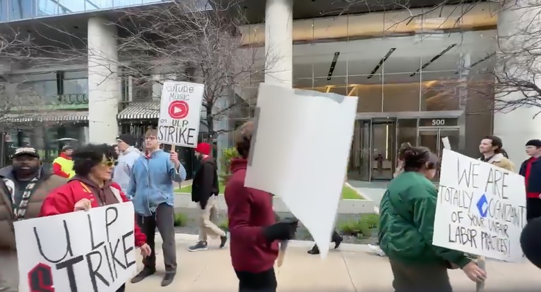 A picket line of strikers protesting outside of Google’s Austin, TX offices. Various people marching outdoors, holding picket signs demanding better treatment.